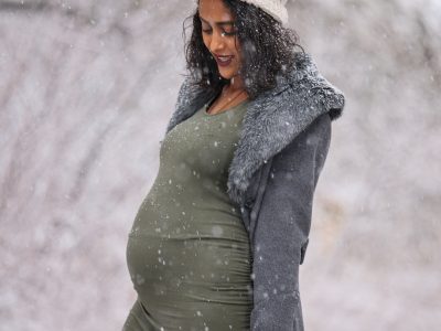 Pregnant woman in a green dress, gray sweater, and cream hat outside in the snow.
