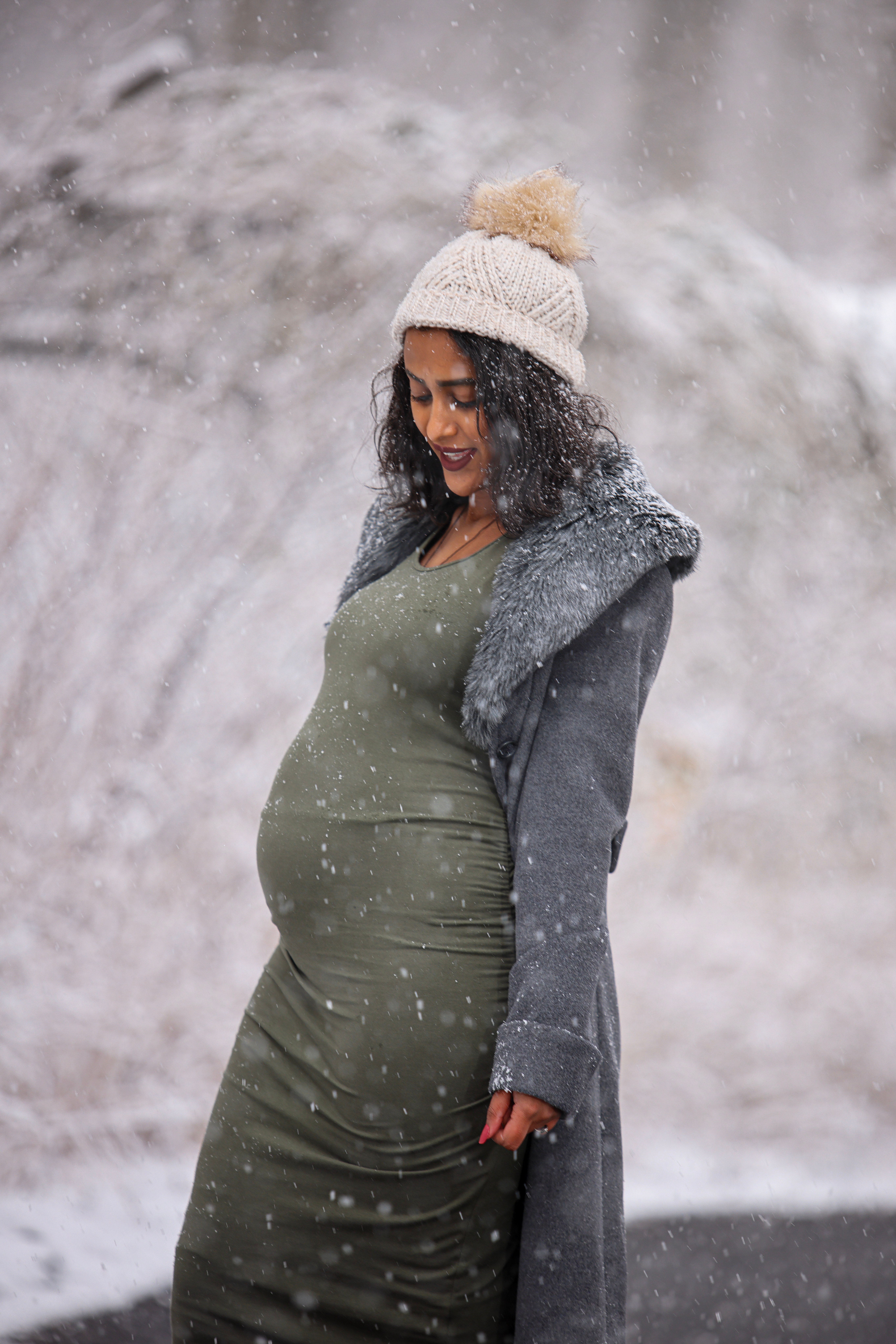 Pregnant woman in a green dress, gray sweater, and cream hat outside in the snow.